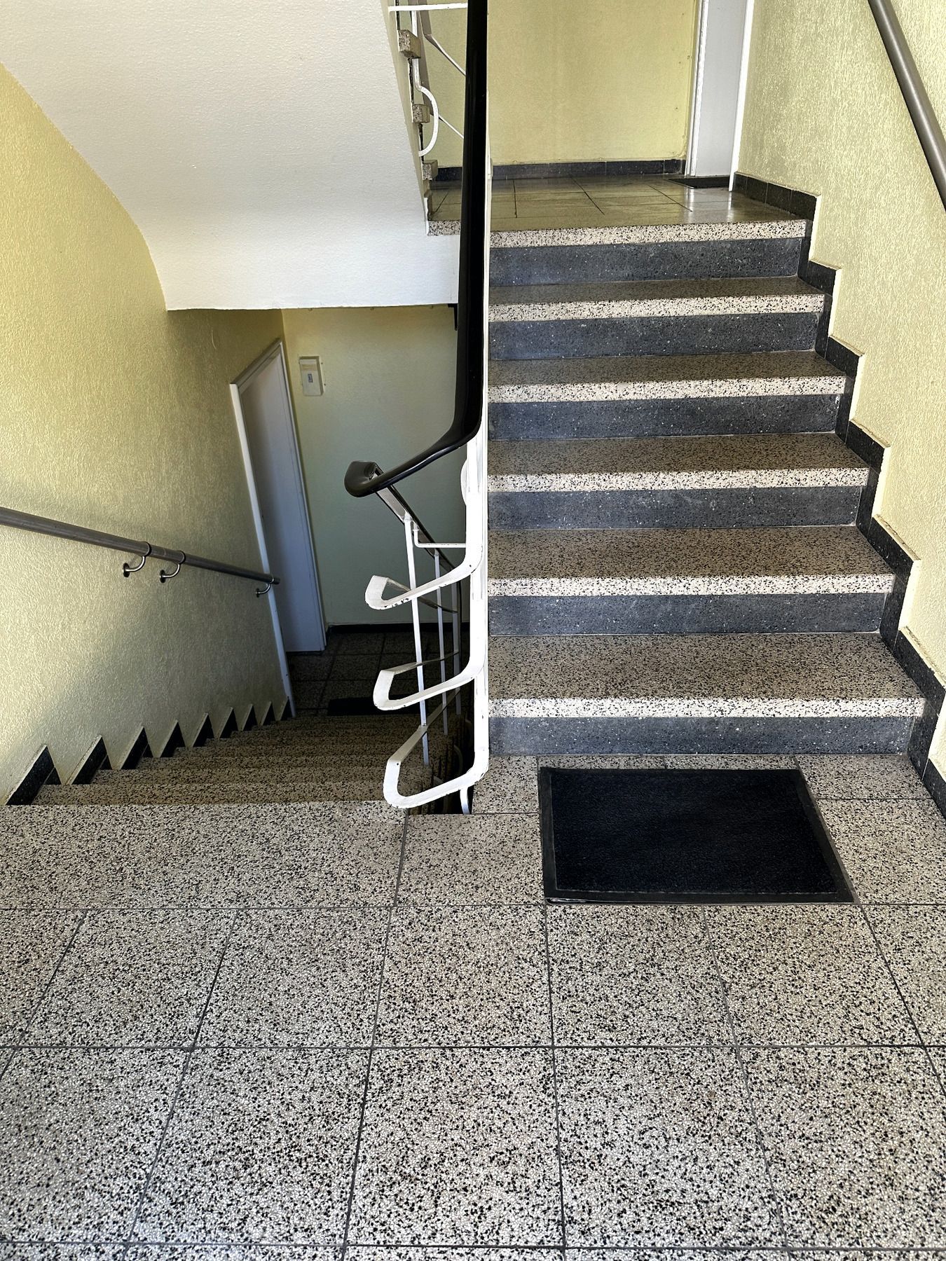 Innentreppenhaus mit grauen Terrazzostufen, weißem Geländer und einer schwarzen Fußmatte am oberen Treppenabsatz.