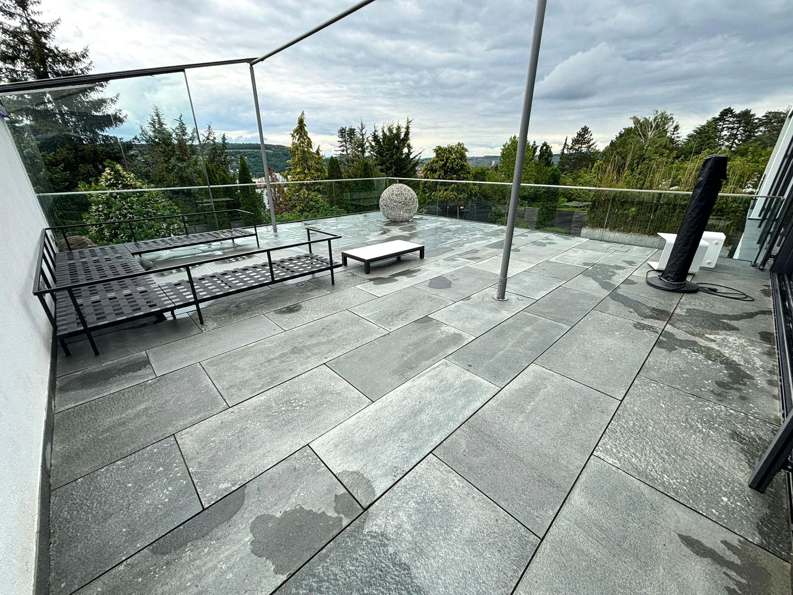 Dachterrasse mit grauen Steinfliesen, Liegestühlen, Topfpflanzen und Blick auf Bäume unter einem Blätterdach.
