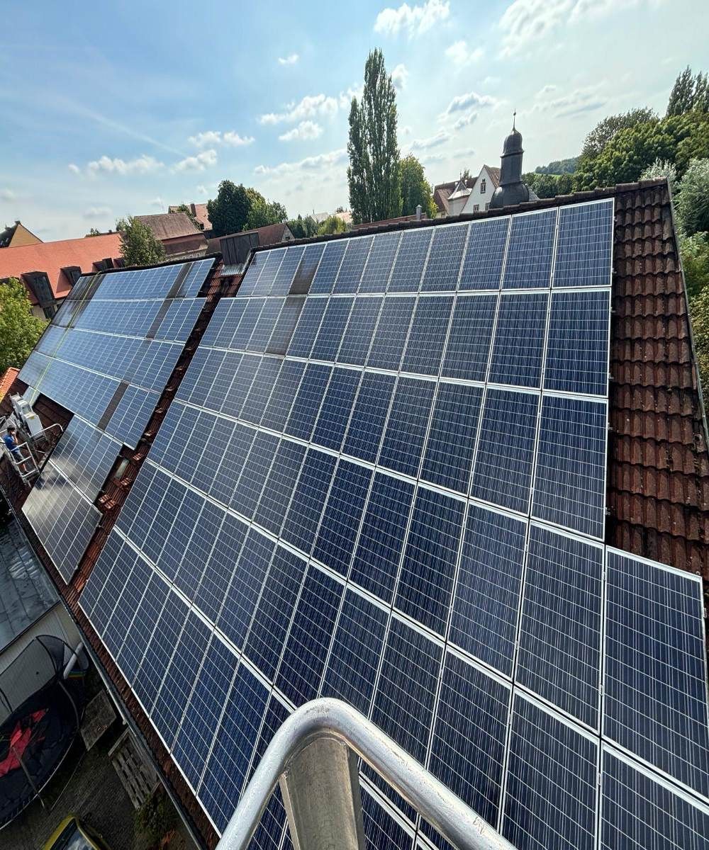 Solarpaneele bedecken ein schräges Dach unter blauem Himmel, im Hintergrund sind Bäume und Häuser zu sehen.