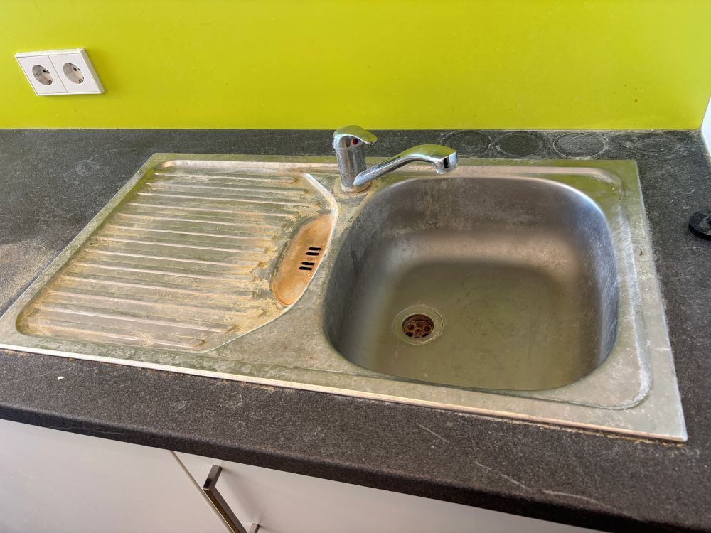 Küchenspüle mit Abtropffläche aus Metall, dunkler Arbeitsplatte und hellgrünem Spritzschutz.