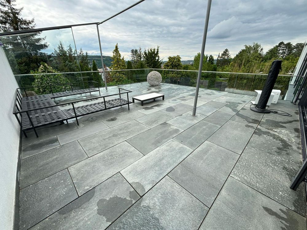Dachterrasse mit grauen Steinfliesen, schwarzen Bänken und einer runden Feuerstelle mit Blick auf Bäume und Wasser.