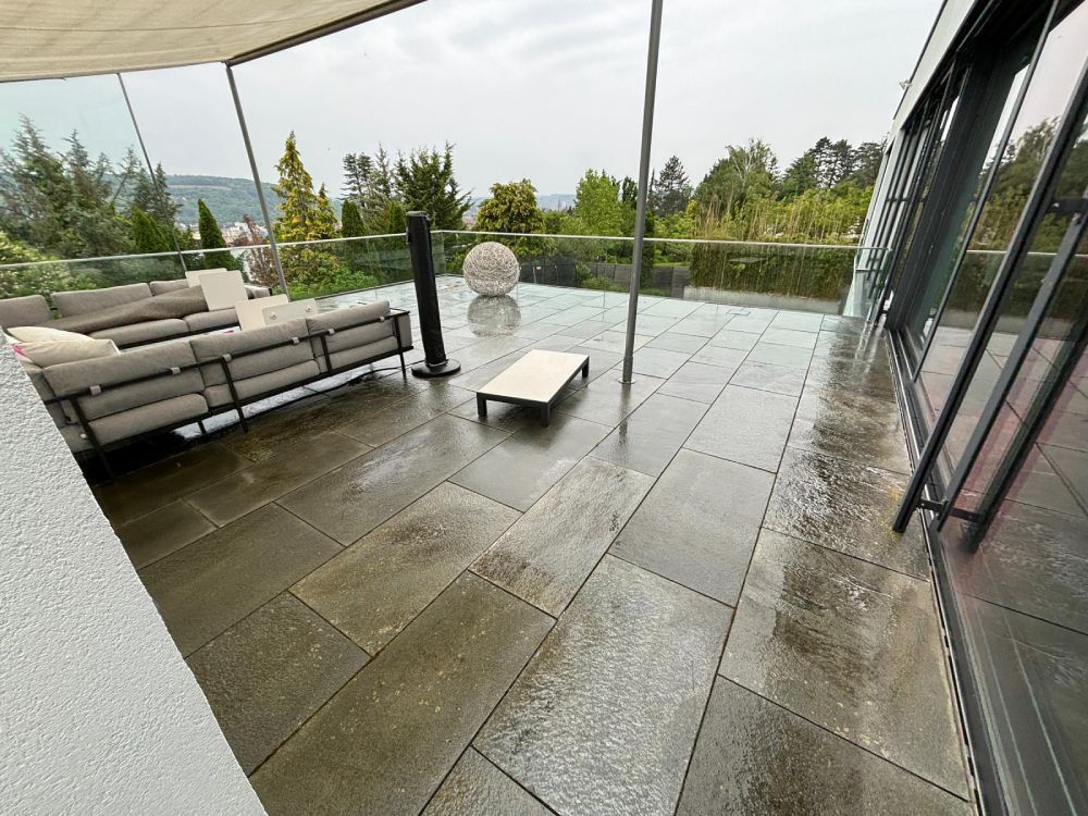 Moderne Terrasse mit grauen Steinfliesen, Gartensofa, niedrigem Tisch und Glasgeländer mit Blick auf die Bäume