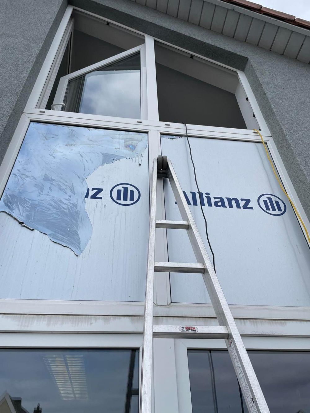 Eine Leiter lehnt an einer Hauswand unter einem hohen Fenster; die Fassade ist weiß verkleidet und mit einer blauen Schutzfolie versehen.