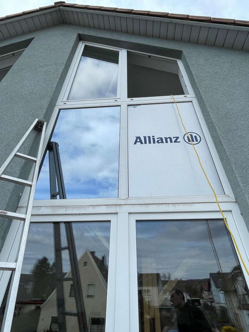 Das Schaufenster der Allianz-Filiale an einem grauen Gebäude, mit Himmelsspiegelungen und einer Leiter daneben