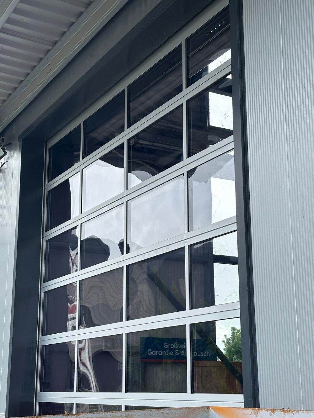 Weiß gerahmtes Fenster mit Spiegelungen und teilweise sichtbarer Innenansicht, von außerhalb eines Wellblechgebäudes betrachtet.