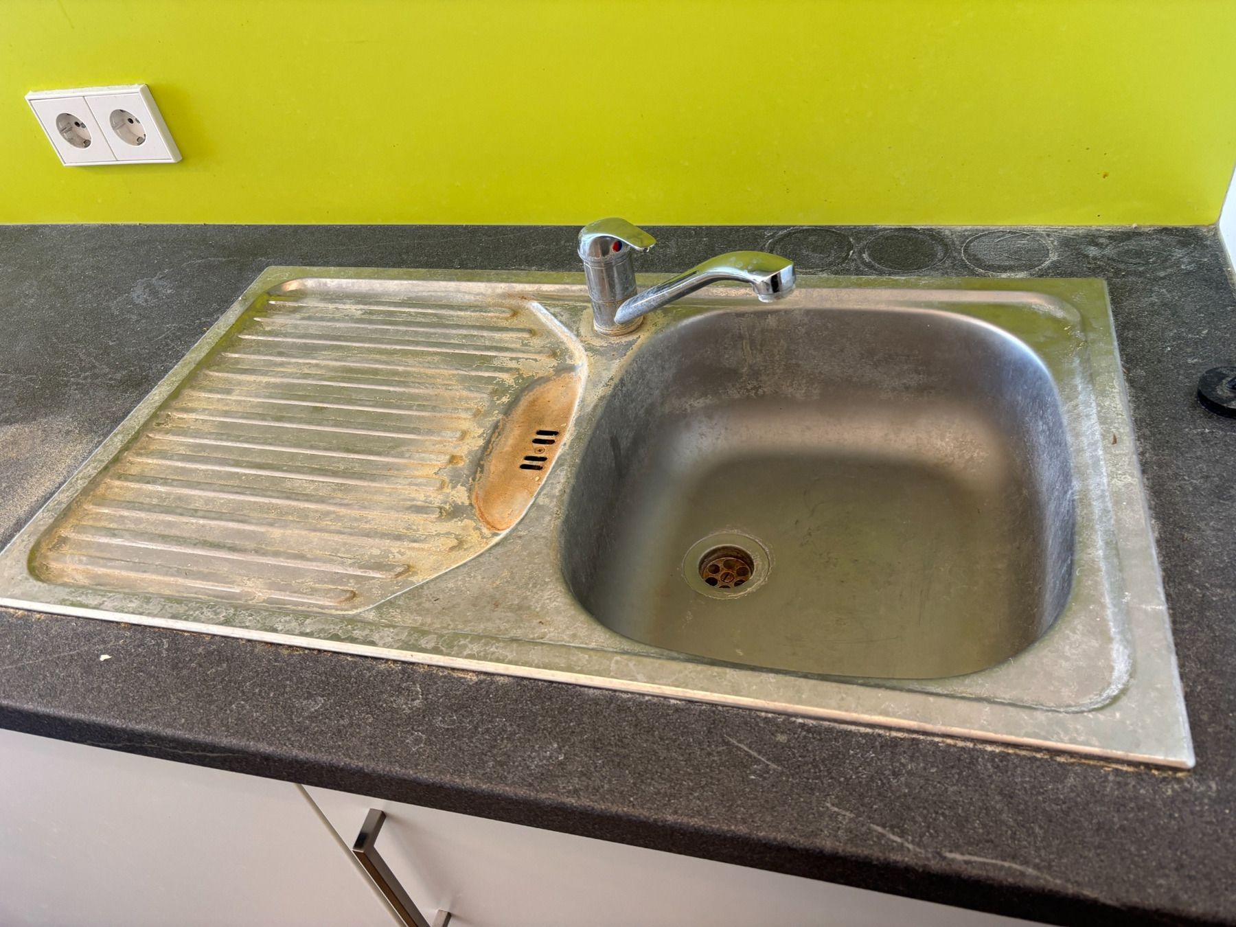 Küchenspüle mit grauer Arbeitsplatte, verchromter Armatur und gelbgrüner Wand dahinter