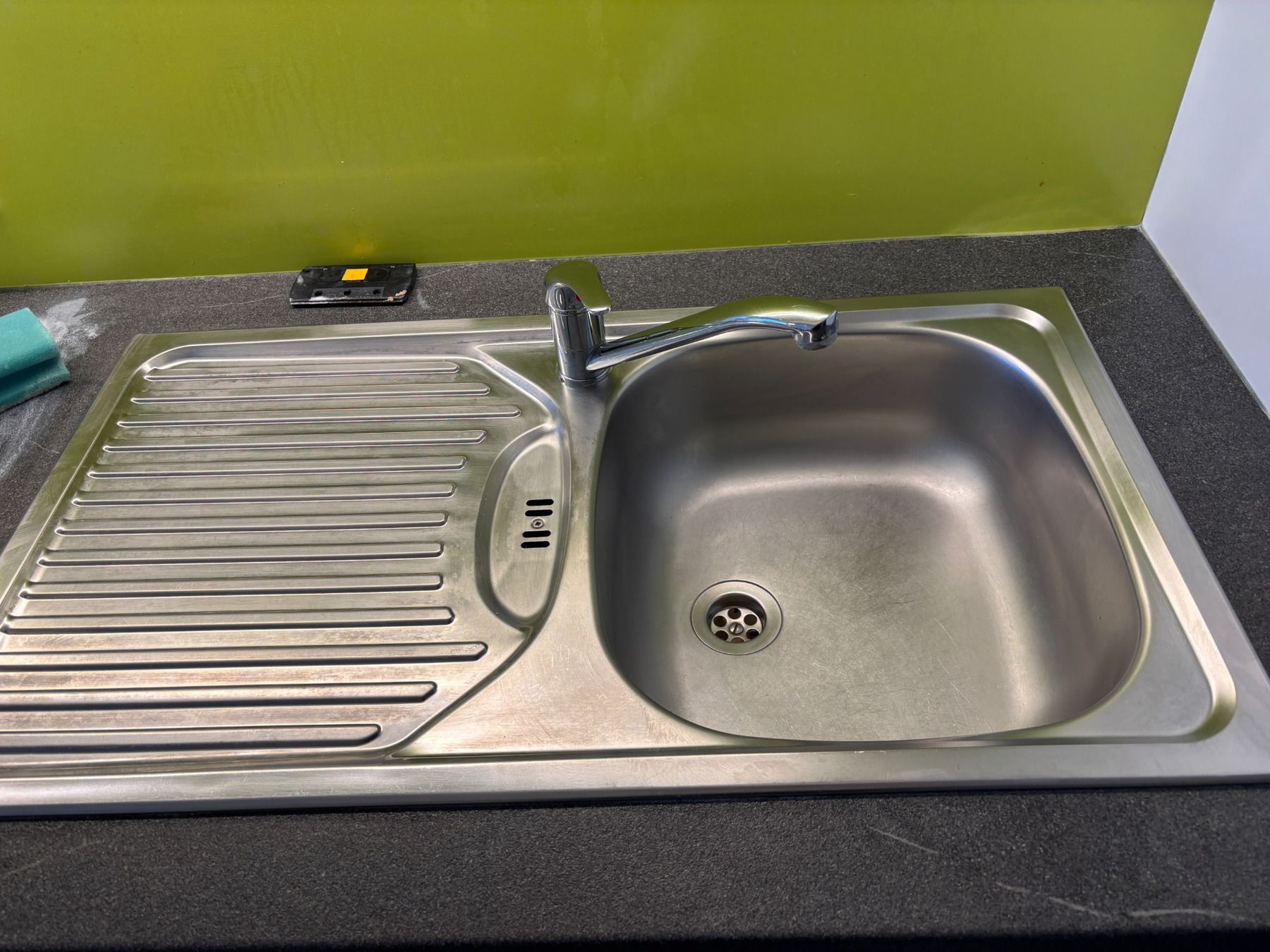 Edelstahl-Küchenspüle mit Abtropffläche neben einer grünen Wand auf einer gesprenkelten Arbeitsplatte.