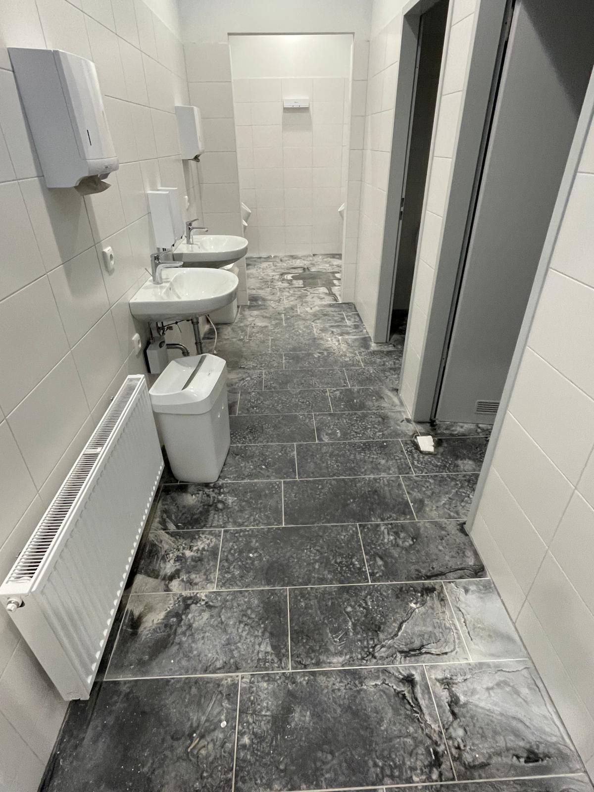 Schmaler öffentlicher Toilettengang mit gefliestem Boden, wandmontierten Waschbecken, Toiletten und offenen Kabinentüren