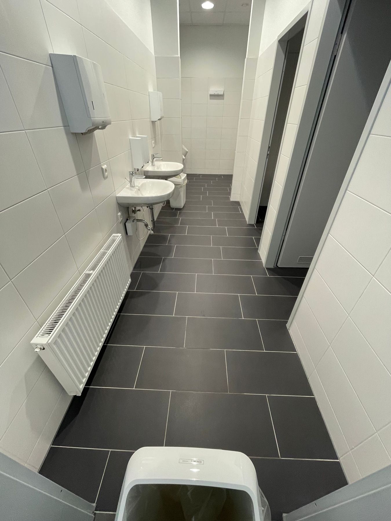 Leerer öffentlicher Toilettenkorridor mit weiß gefliesten Wänden, dunklen Bodenfliesen, Waschbecken, Heizkörpern und Kabinentrennwänden.