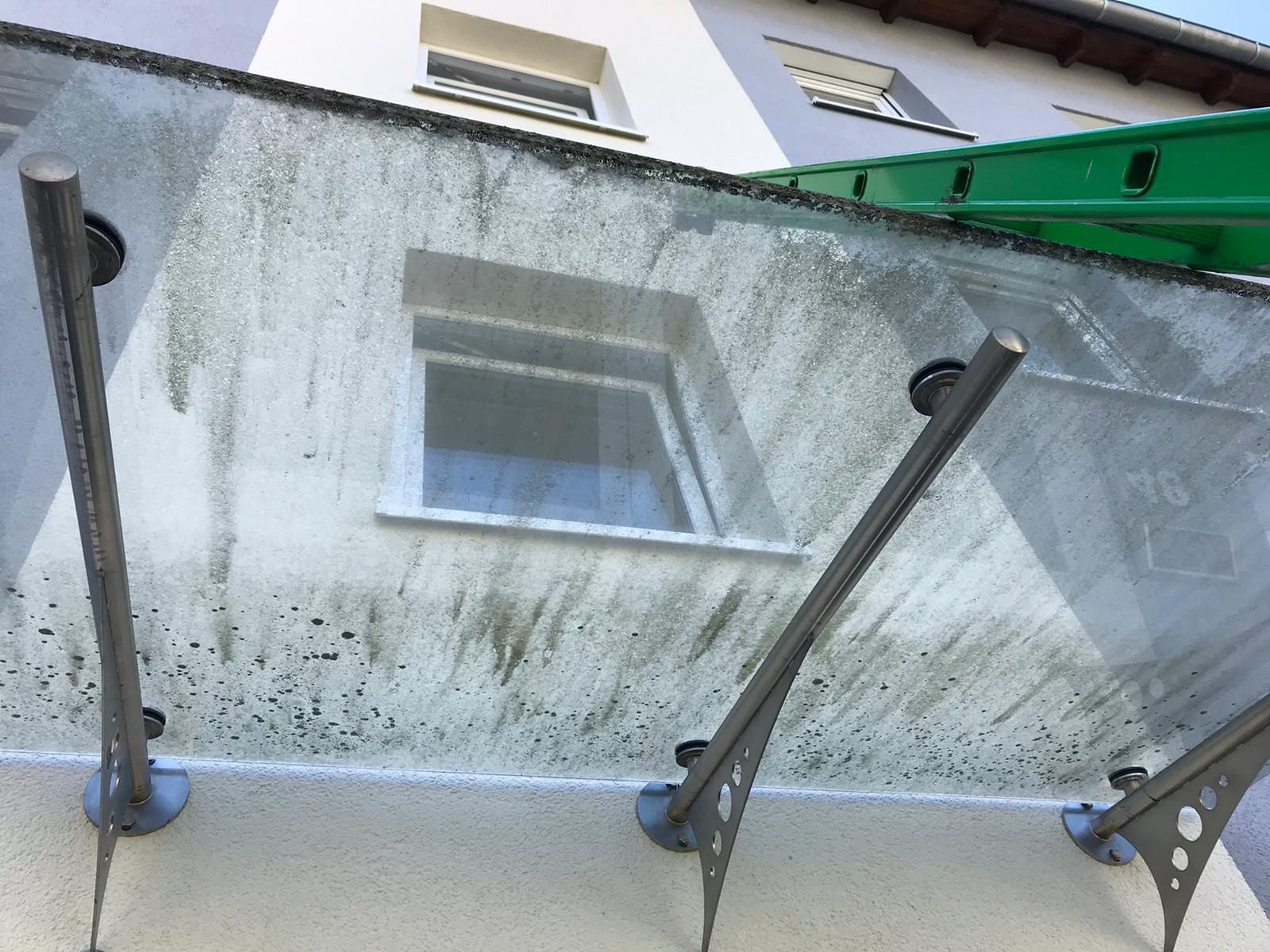Blick nach oben auf eine Gebäudefassade hinter einem gläsernen Balkongeländer mit Metallstützen und grüner Markise.