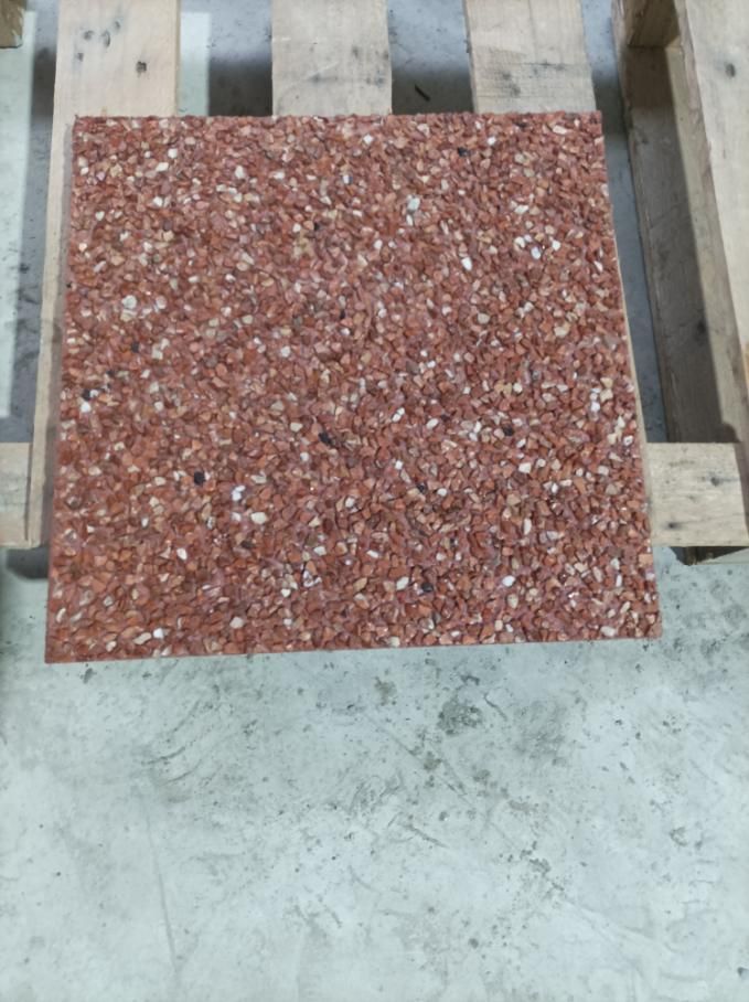 Azulejo cuadrado de piedra moteada de color rojo sobre una paleta de madera, que reposa sobre un piso de hormigón gris.