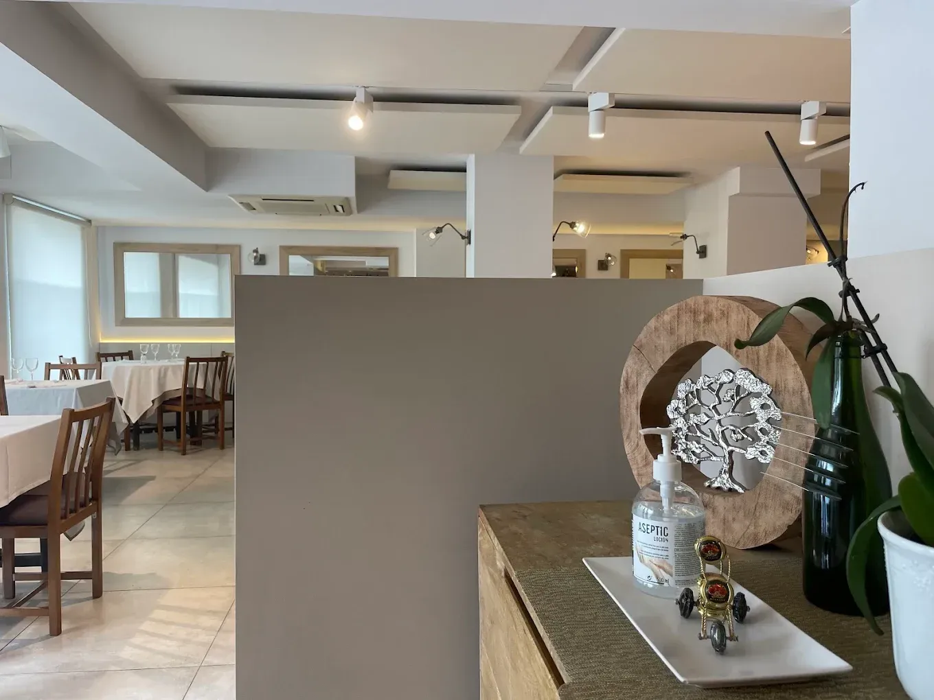 Interior luminoso de una cafetería con mesas de madera, paredes blancas y una vitrina decorativa en el mostrador.