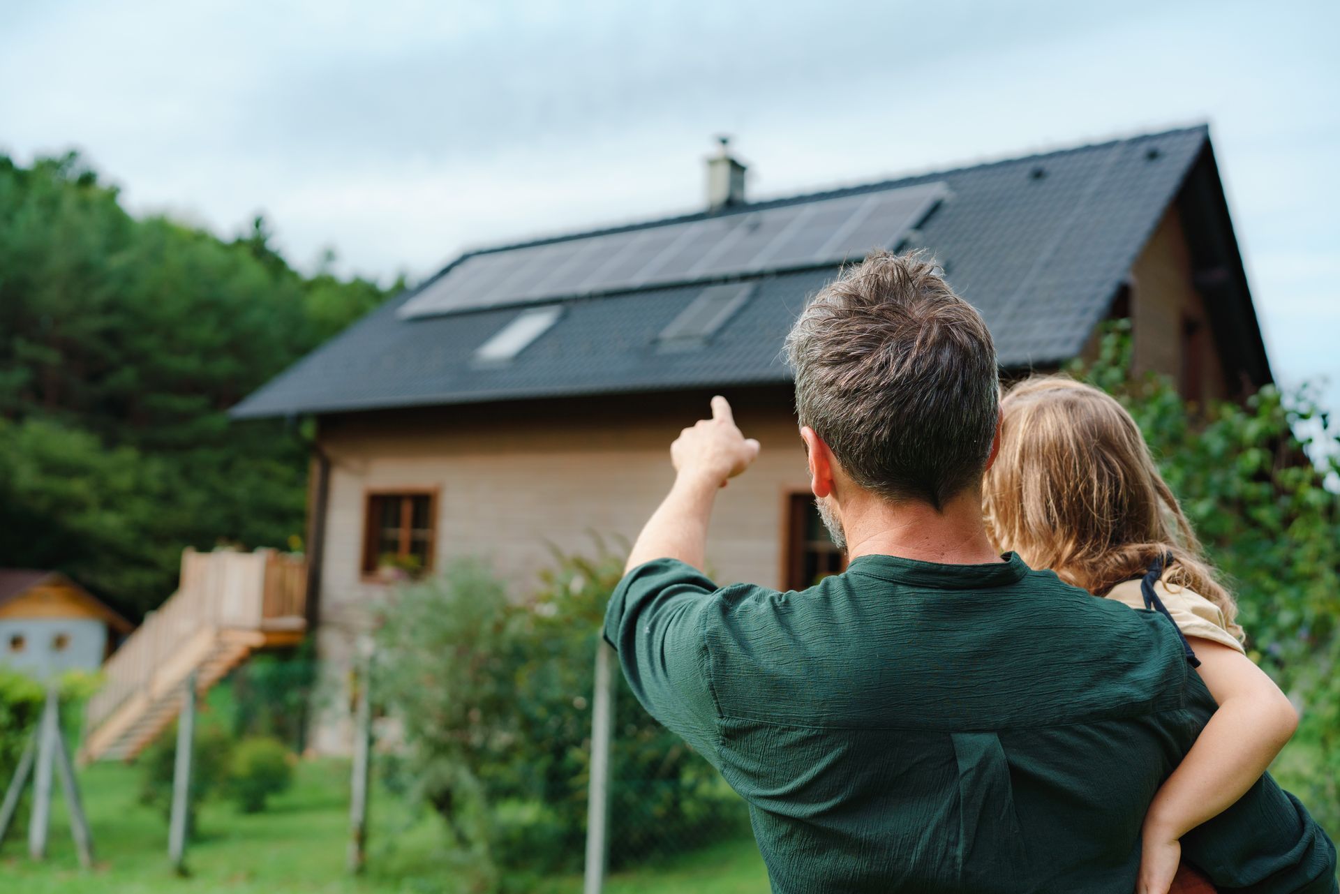 Une personne désigne une maison en tenant un enfant dans ses bras, avec un jardin verdoyant et des arbres à proximité.