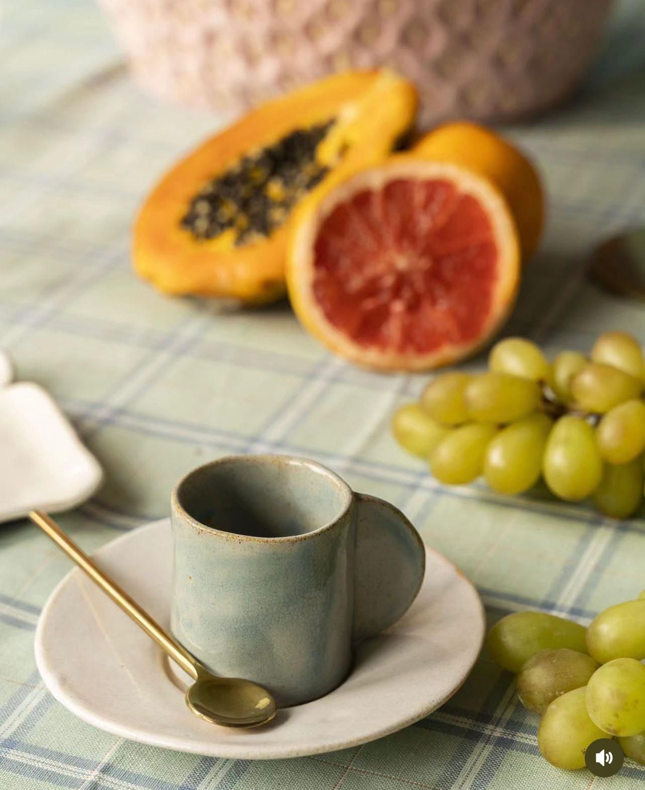 Una taza y un platillo con una cuchara sobre una mesa con fruta al fondo.
