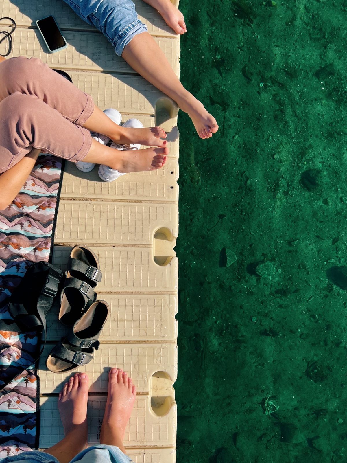 Gente sentada en un muelle con los pies colgando sobre el agua verde; sandalias y un teléfono descansan cerca.