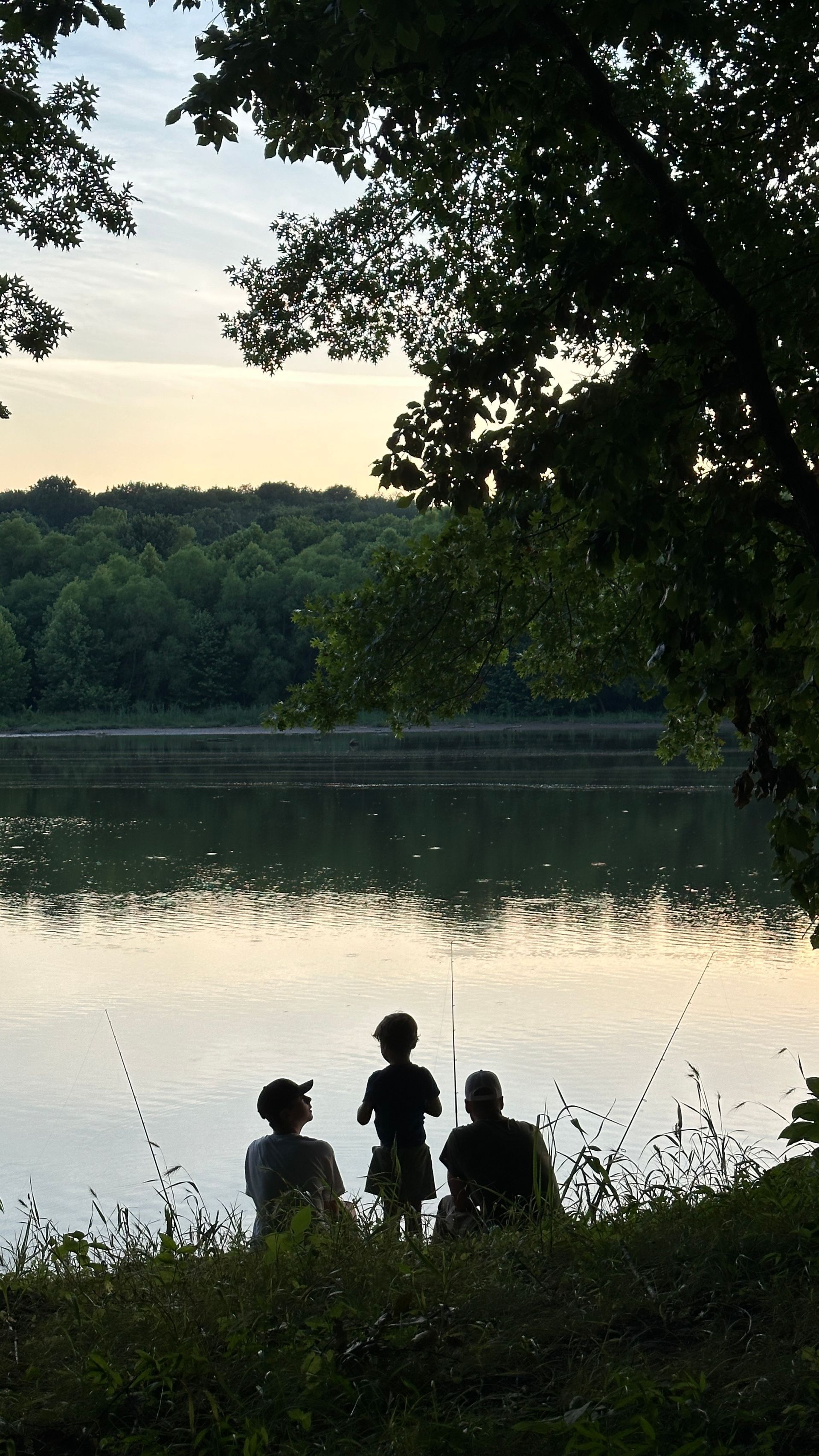Tres personas pescando junto a un lago al anochecer, recortadas contra el agua y los árboles.