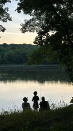 Tres personas pescando junto a un lago al anochecer, recortadas contra el agua y los árboles.