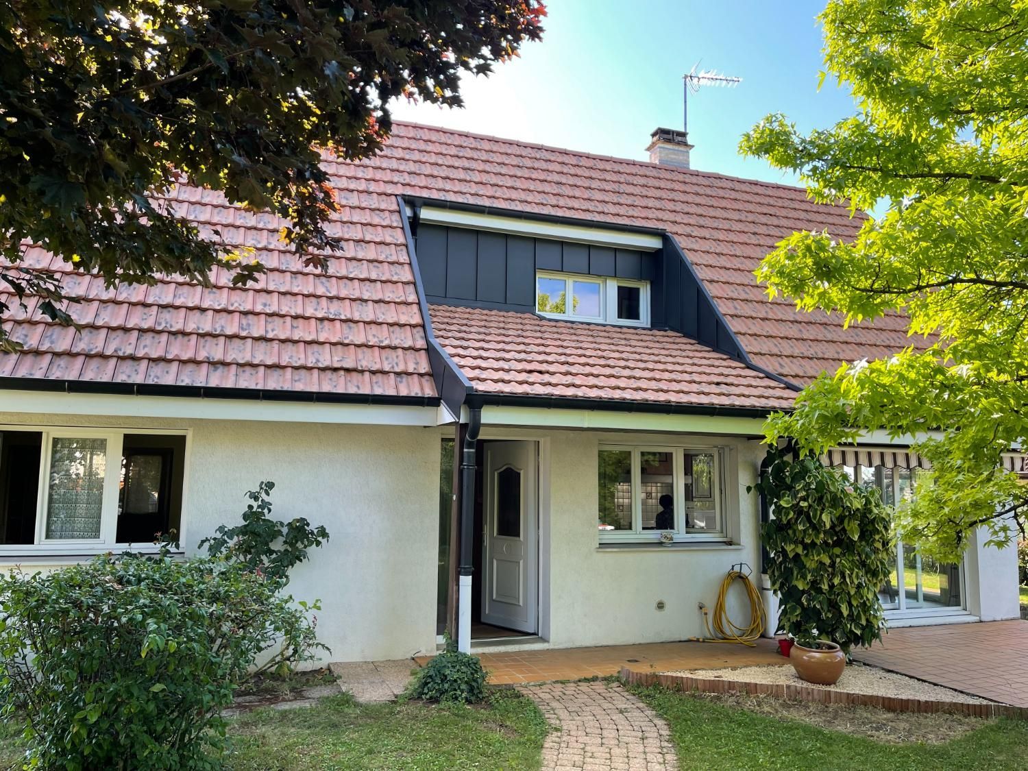 Maisonnette au toit de tuiles rouges, aux murs clairs et avec une petite cour avant arborée.