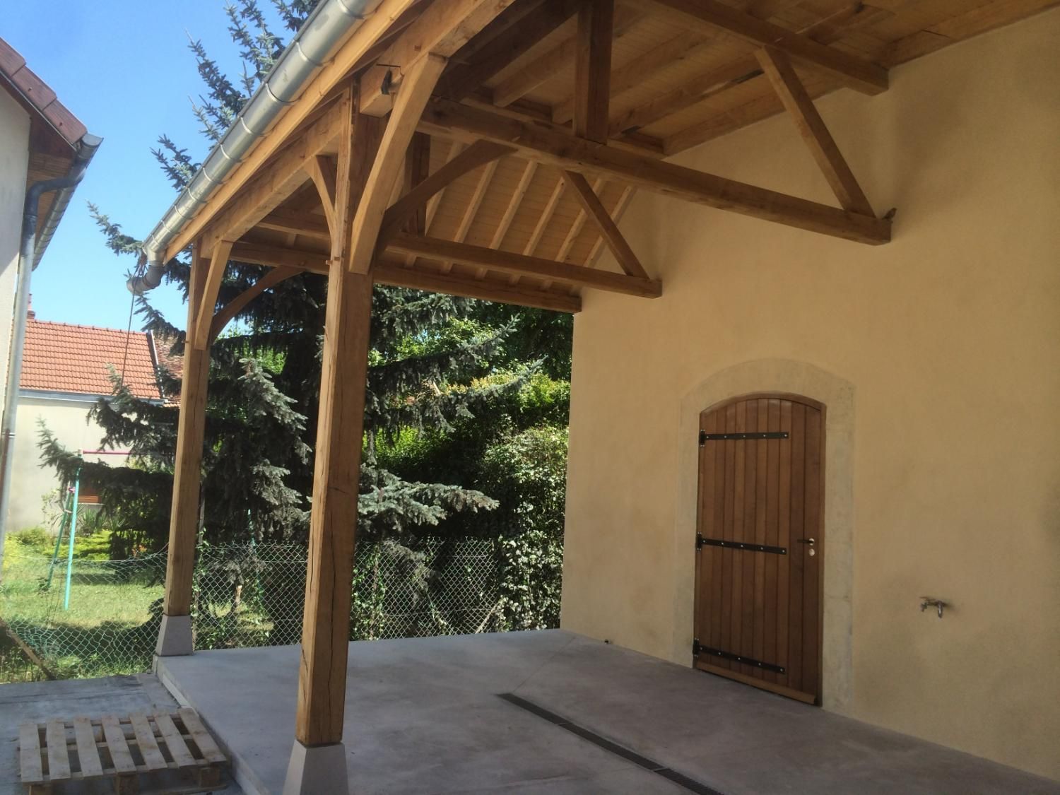 Porche en bois avec un toit clair et des poutres de soutien, jouxtant un bâtiment avec une porte en bois.