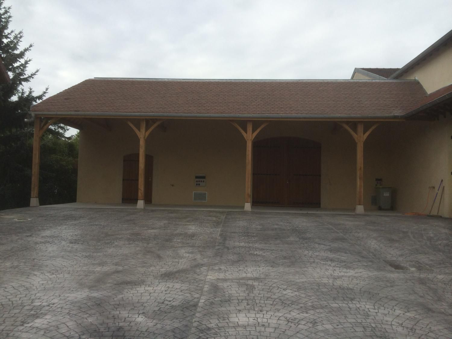 Abri voiture avec supports en bois et toit marron ; allée carrossable devant.