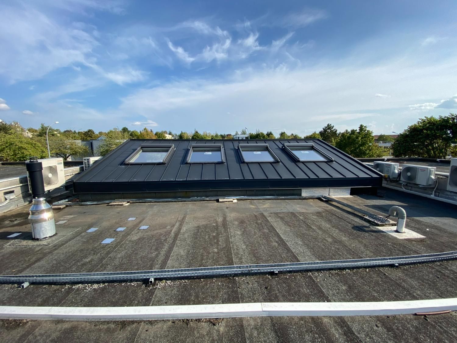 Un toit avec une partie surélevée en métal noir percée de puits de lumière, par une journée de ciel bleu.