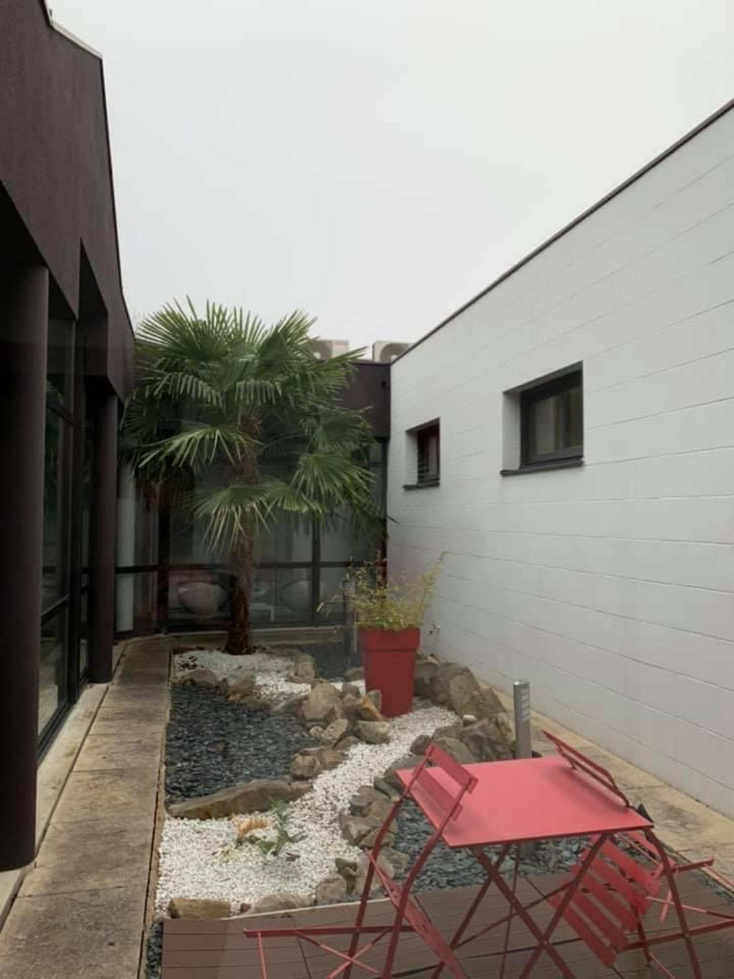 Cour intérieure avec palmier, chaise rouge et jardinière rouge entre des bâtiments blancs et sombres.