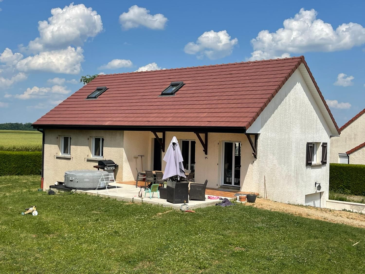 Maison beige avec toit rouge et ciel bleu. Mobilier de terrasse, jacuzzi et jardin à l'avant.