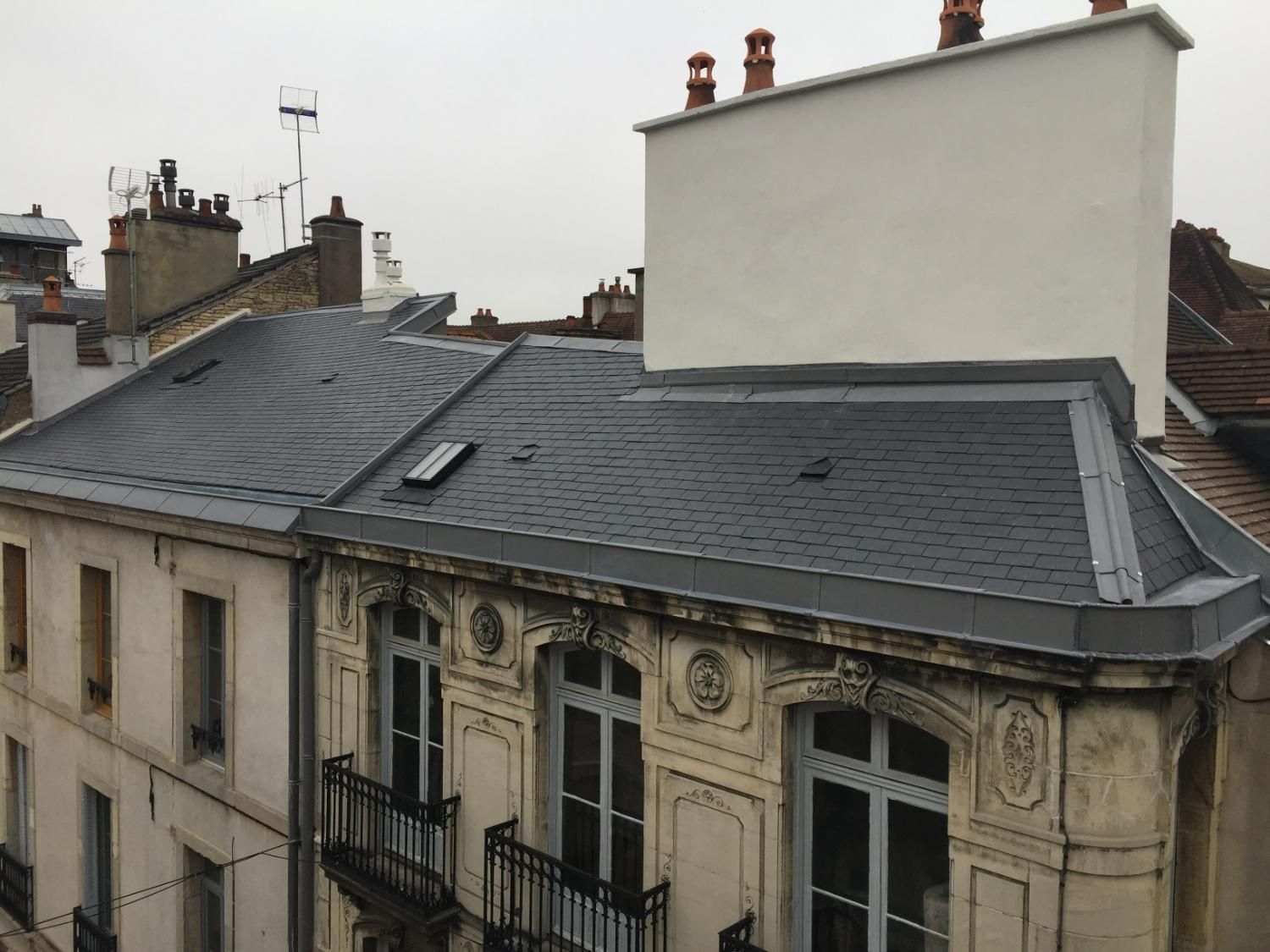 Toits de bâtiments européens aux bardeaux gris foncé, murs blancs, cheminées et ciel couvert.