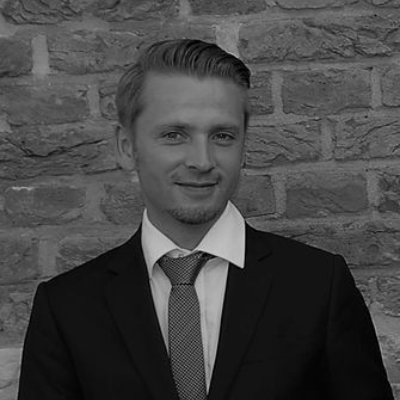 Mann in Anzug und Krawatte lächelt vor einer Backsteinmauer.