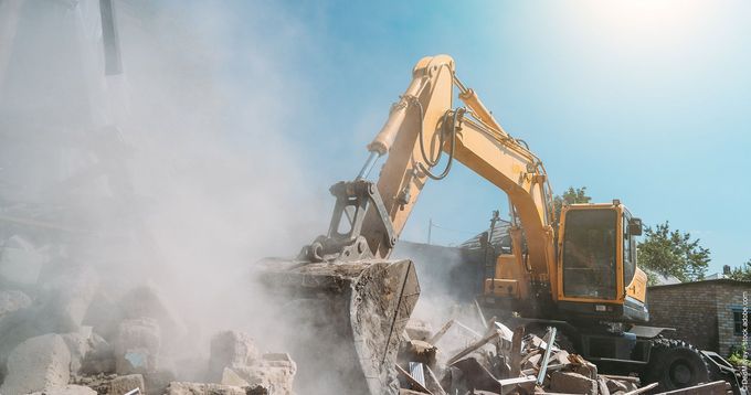 Ein gelber Bagger reißt ein Gebäude ab und wirbelt Staub vor dem blauen Himmel auf.