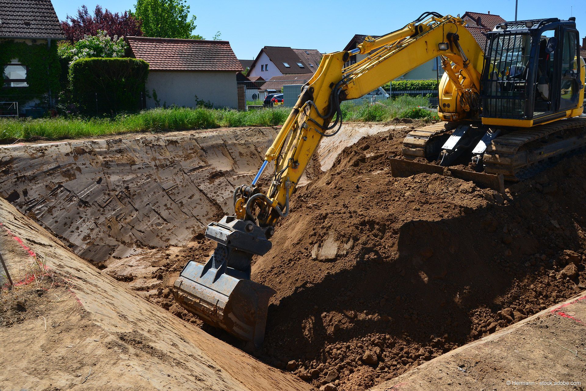Ein gelber Bagger bei Erdarbeiten in einer Wohnsiedlung hebt Erdreich aus einer tiefen Baugrube.