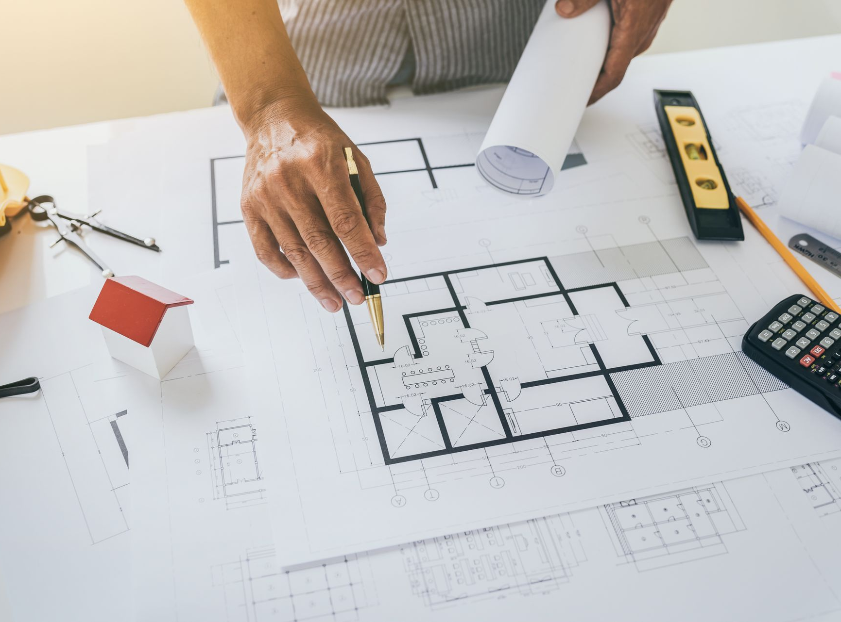 Des mains traçant un plan d'étage sur des plans architecturaux à l'aide d'une règle, d'une calculatrice et de plans enroulés à proximité