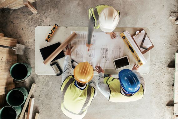 Des ouvriers du bâtiment, casqués, examinent des plans autour d'une table sur un chantier.