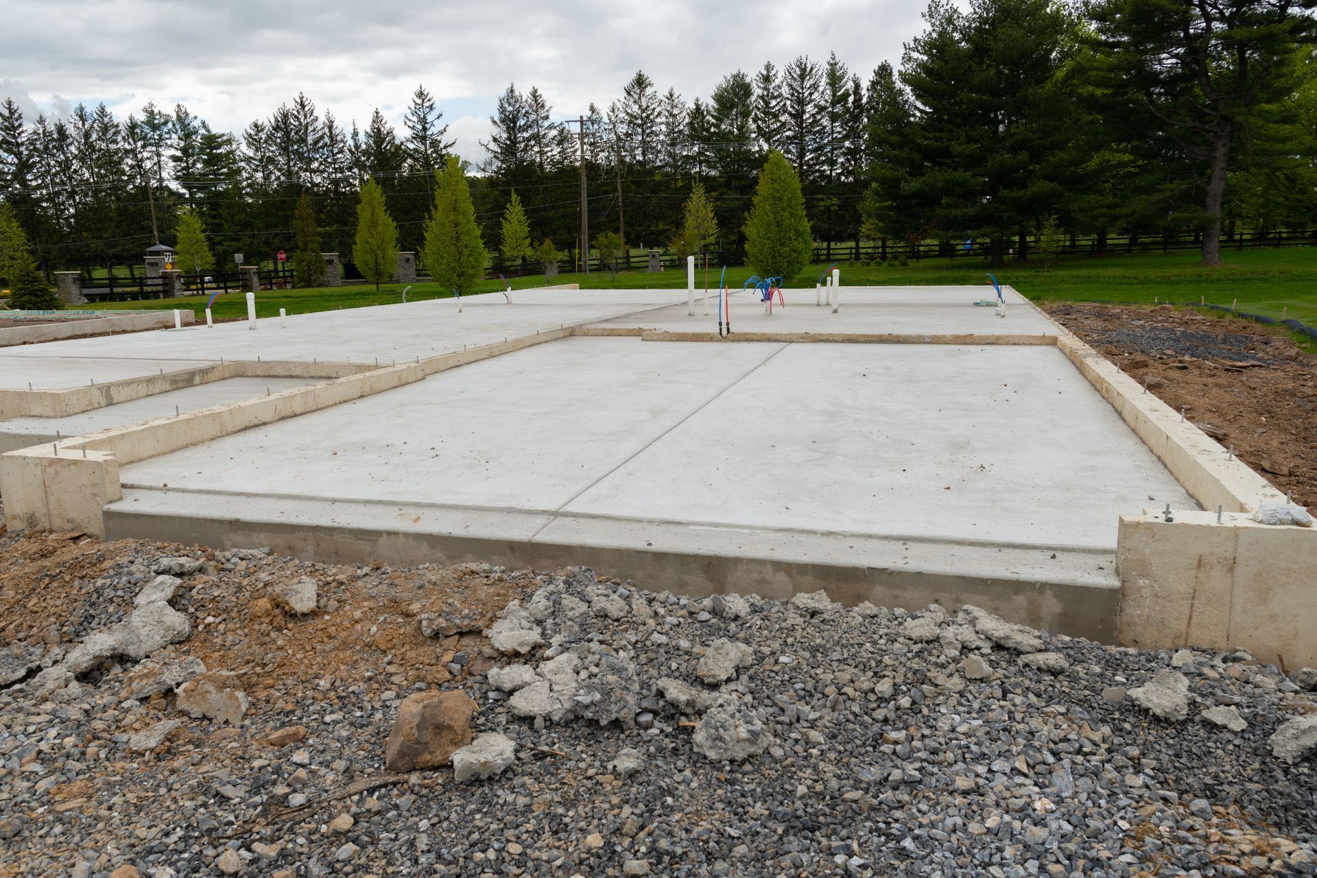 Dalle de fondation en béton sur un chantier de construction, avec du gravier au premier plan et des arbres en arrière-plan.