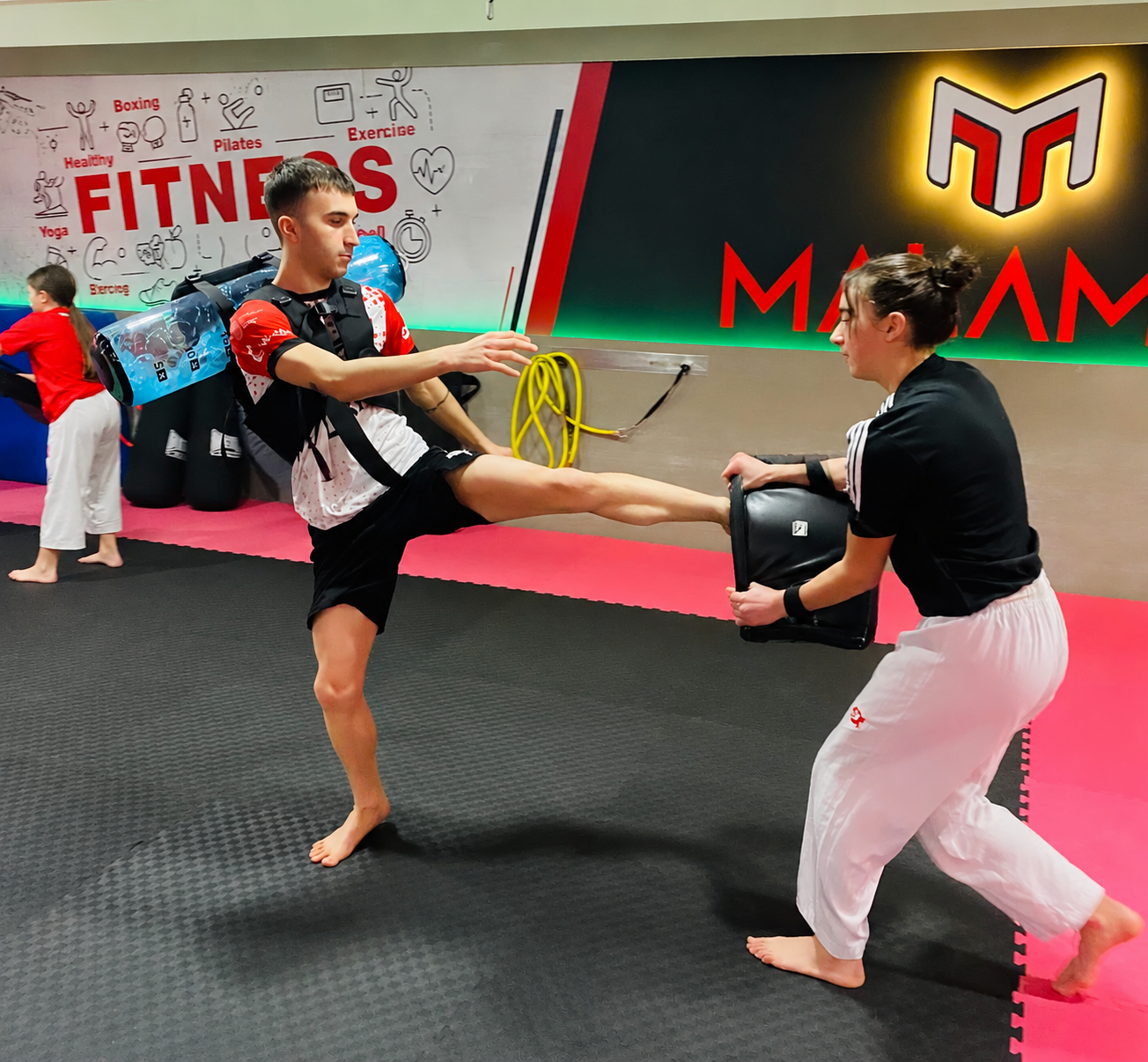 Dos personas practicando patadas de artes marciales en un gimnasio; una sostiene una almohadilla y la otra golpea.
