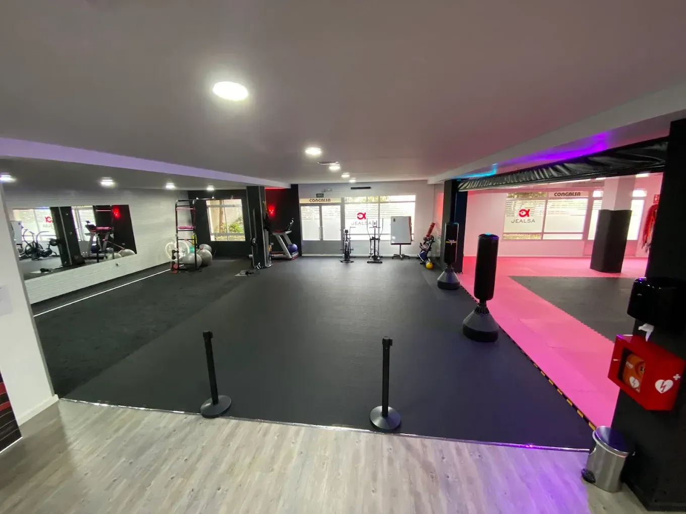 Gimnasio vacío con colchonetas negras, postes de barrera, espejos e iluminación ambiental rosa.