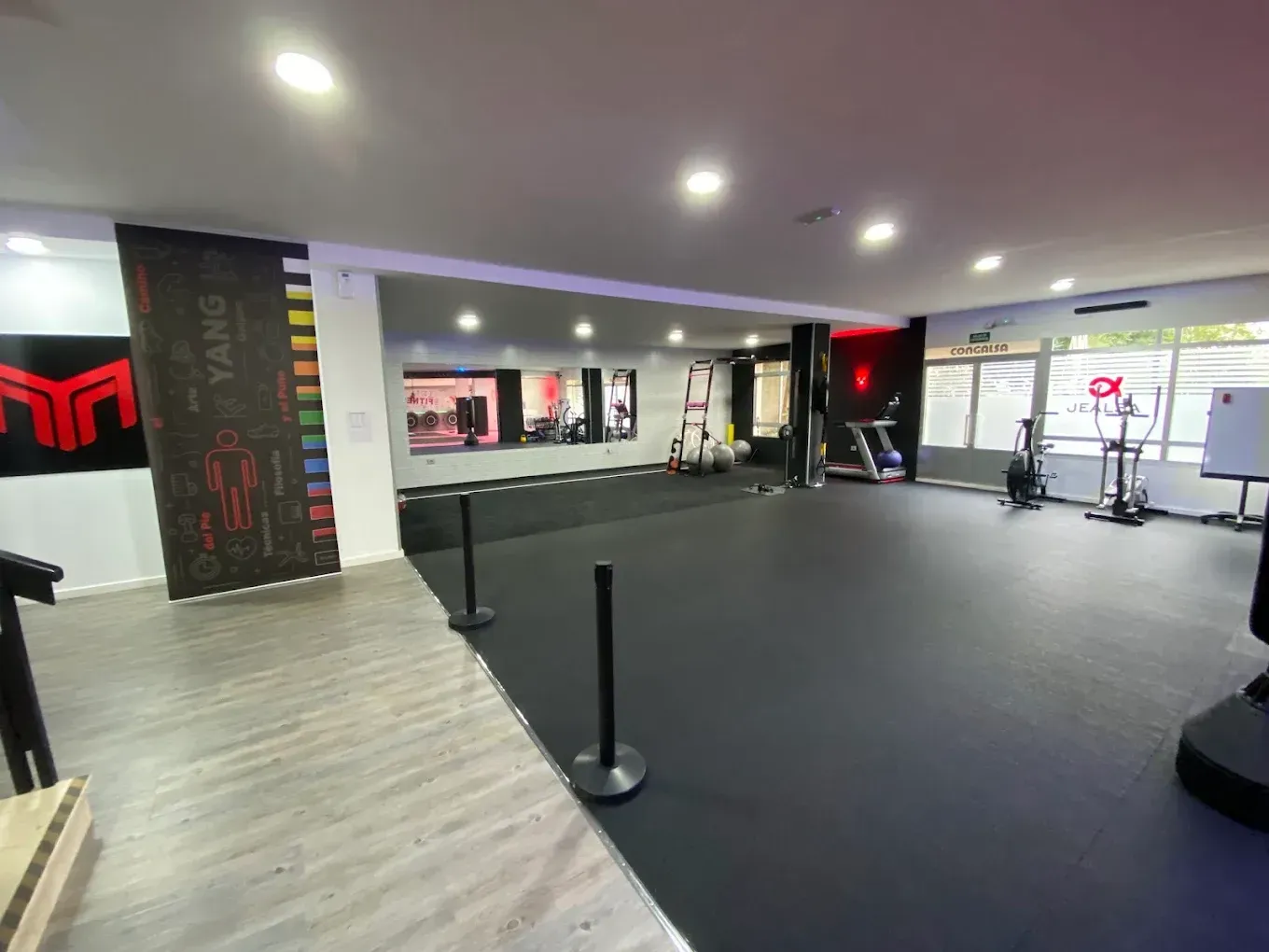 Amplio interior de gimnasio con suelo negro, espejos, mancuernas y equipo cardiovascular bajo luces empotradas.