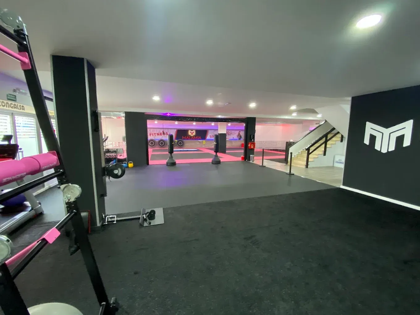 Interior de gimnasio moderno con suelo negro, iluminación rosa, equipamiento y escaleras que conducen a un nivel superior.