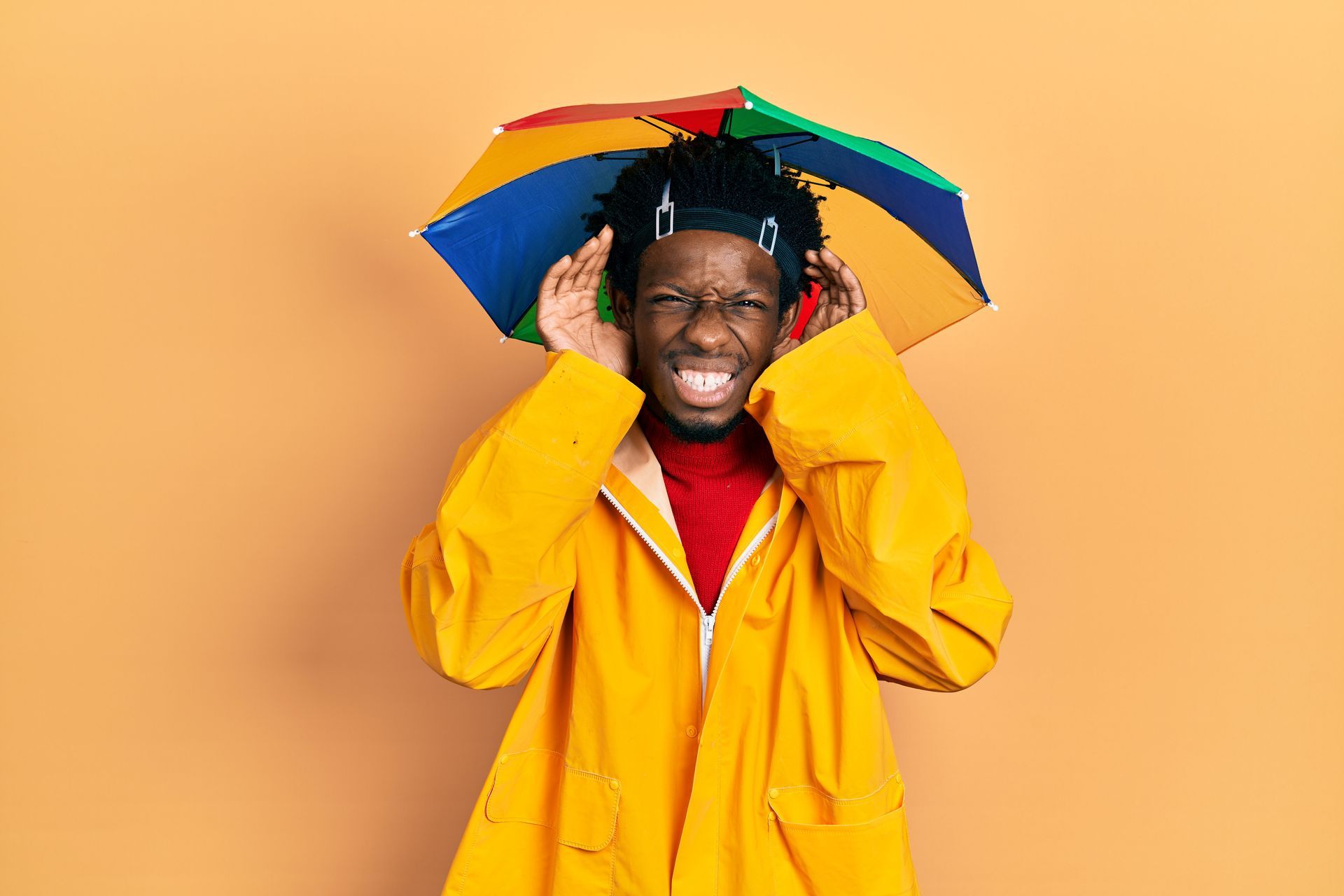 Un homme avec un chapeau-parapluie et un ciré jaune tend les deux oreilles en faisant la grimace