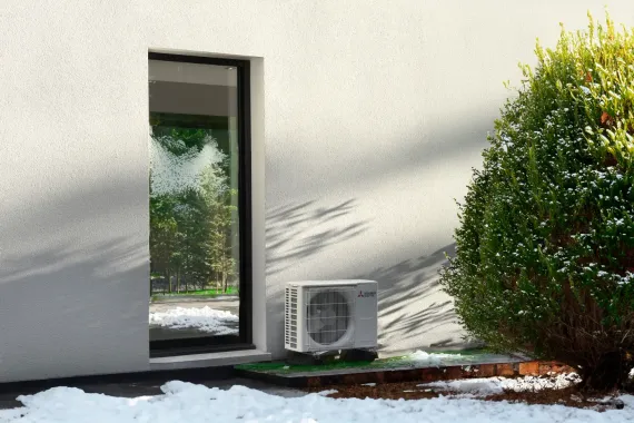 Mur blanc avec une porte vitrée étroite, une unité de climatisation extérieure et des arbustes à côté d’une plaque de neige.