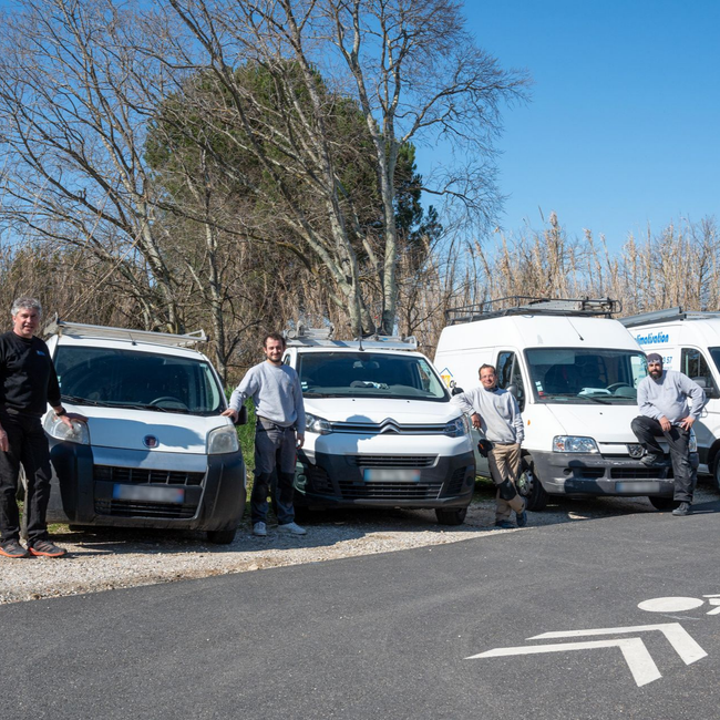 Quatre personnes se tiennent à côté de quatre camionnettes et voitures blanches garées au bord de la route par une journée ensoleillée.