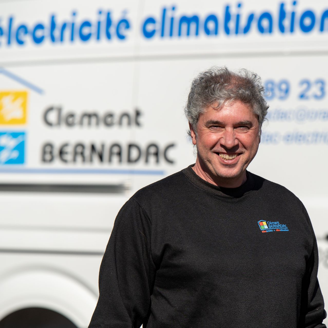 Technicien souriant en pull noir, debout devant un utilitaire blanc portant l'enseigne de l'entreprise