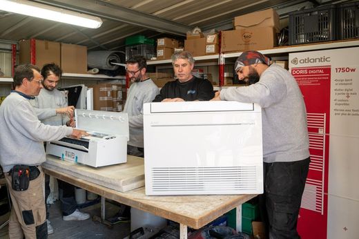 Quatre techniciens déballent du matériel dans un atelier, avec une grande unité de climatisation sur un établi.