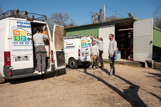 Des ouvriers chargent des fournitures dans des fourgonnettes blanches devant un hangar, dans une cour de campagne ensoleillée