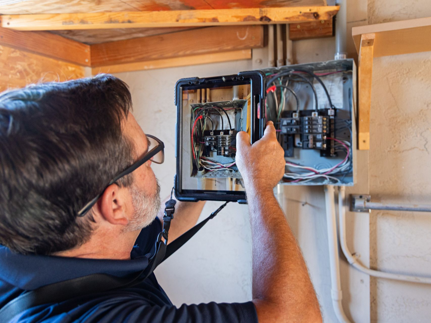Un électricien inspecte un tableau électrique ouvert, avec des fils apparents, dans un mur de maison.