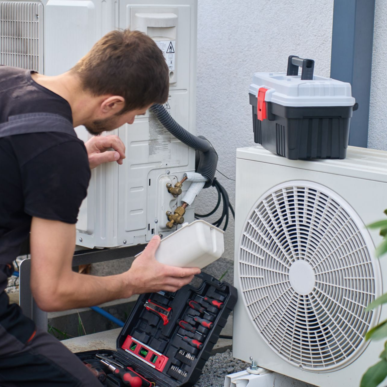Technicien réparant une unité de climatisation extérieure à côté du panneau de service ouvert, avec des outils et un récipient blanc