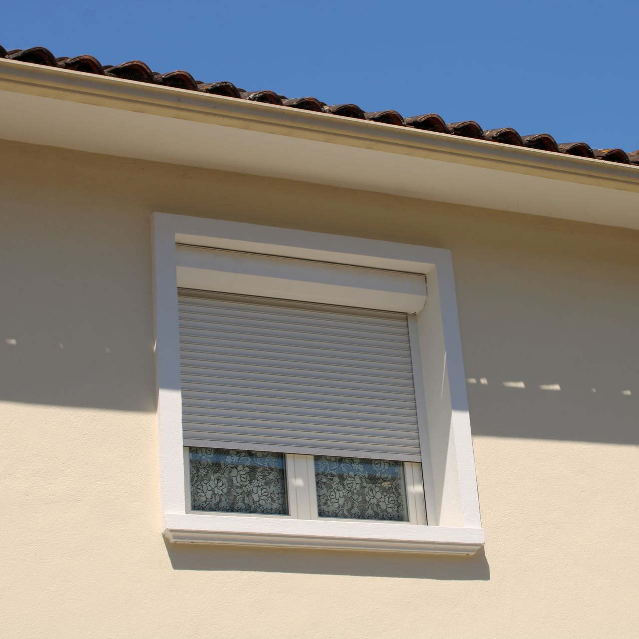 Fenêtre d’une maison blanche avec un volet roulant fermé et des rideaux en dentelle sous un toit de tuiles
