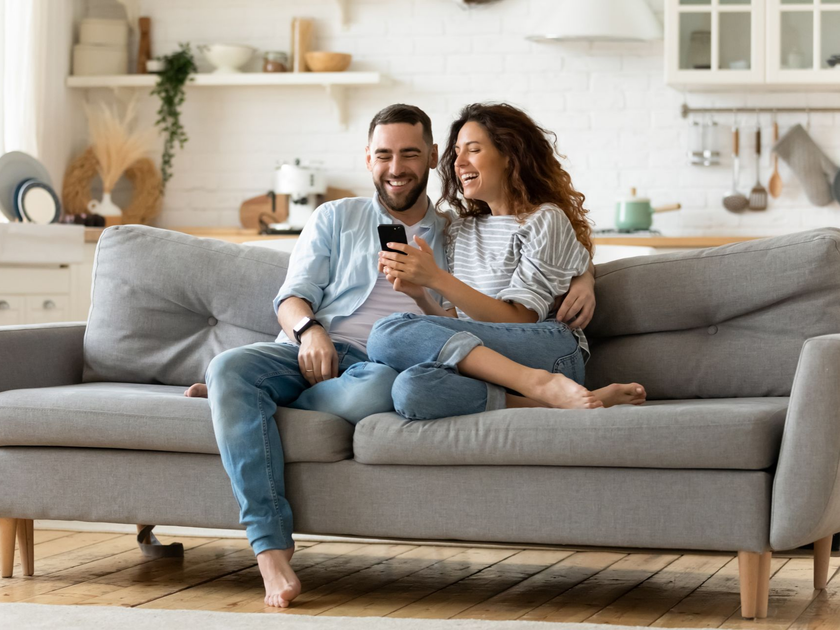 Un couple souriant assis sur un canapé gris, regardant un smartphone dans un salon lumineux.