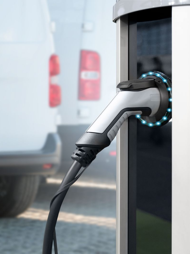 Prise de recharge pour véhicule électrique branchée à une borne de recharge, avec une camionnette blanche en arrière-plan.