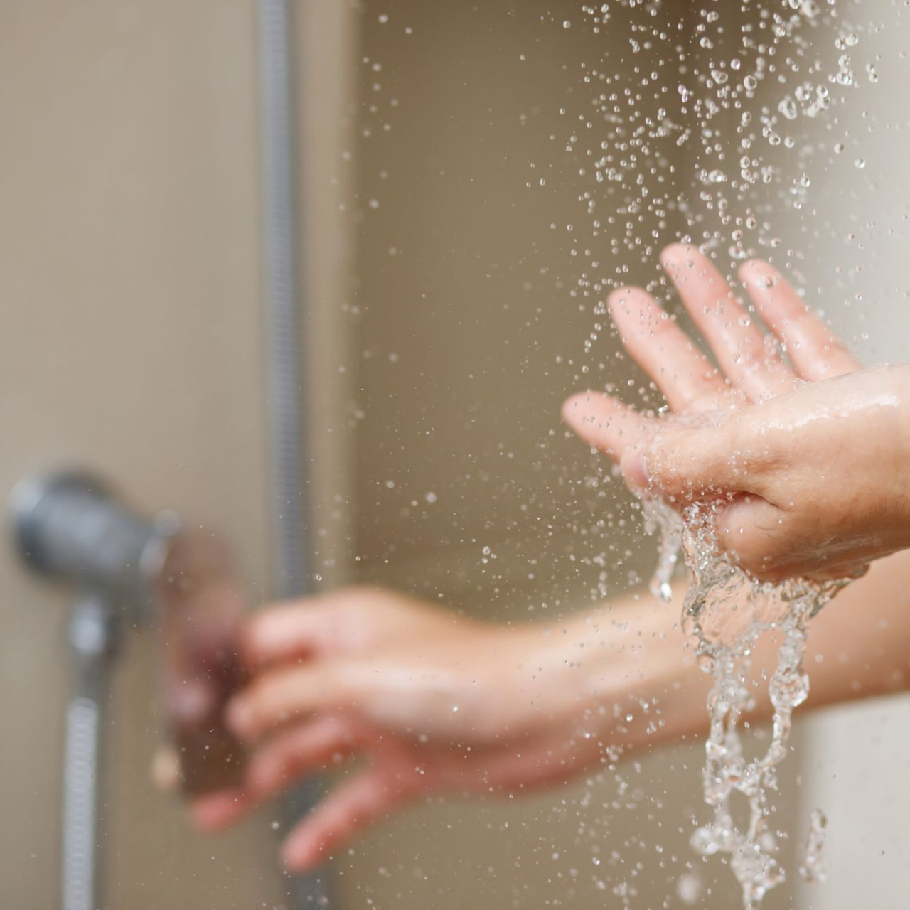 Des mains se rincent sous un jet de douche, tandis que l’eau éclabousse un mur carrelé.